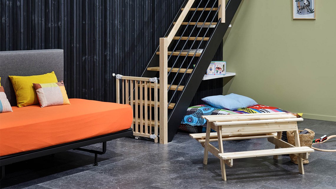 Un espace sous un escalier avec un lit orange et un petit banc en bois. Le mur derrière l'escalier est noir, contrastant avec le mur vert pâle à droite. Des coussins colorés sont disposés sur le lit.